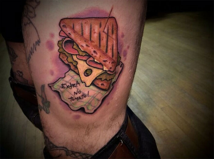Food tattoo of a sandwich with text "Extra no tomato" on a leg, showcasing vibrant colors and bold outlines.
