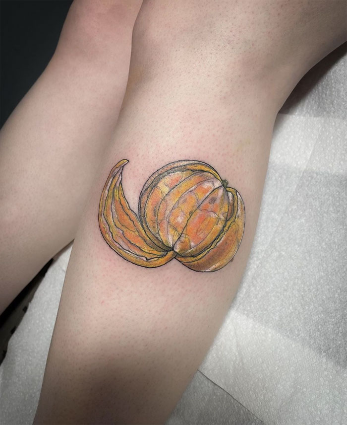 Food tattoo of a vibrant, half-peeled orange with artistic shading on a person's leg.
