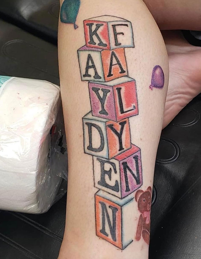 Tattoo fail of misspelled name on child-themed blocks with balloons and teddy bear on lower leg showcasing epic tattoo fails.