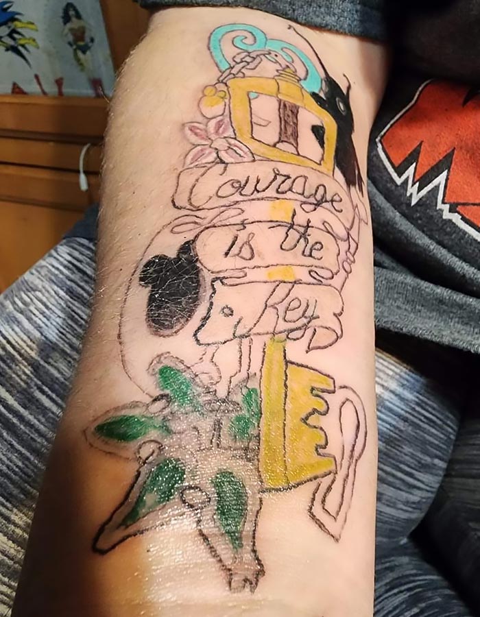 Tattoo fail of a yellow key with a banner reading courage is the key, uneven lines and distorted design on forearm.