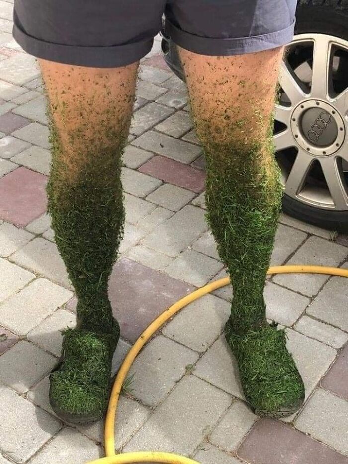 Legs covered in grass clippings, wearing shorts and shoes, standing on a tiled floor next to a hose.