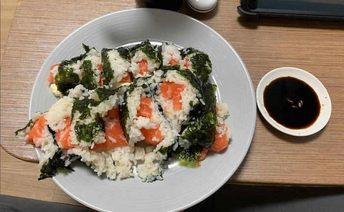 Unappetizing sushi rolls with salmon and rice, falling apart on a plate, served with a small dish of soy sauce.