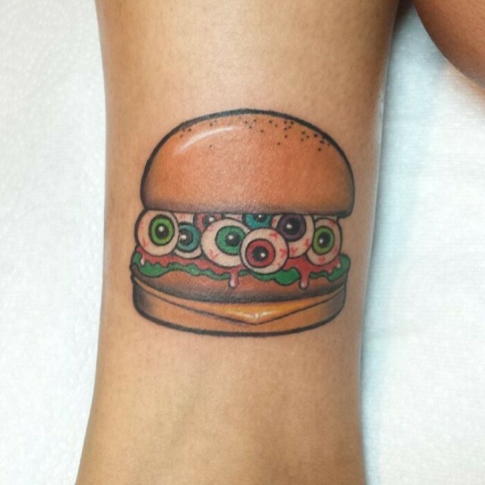 Food tattoo of a burger with colorful eyeballs on arm.