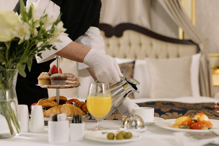 Luxury breakfast in a hotel room with a server pouring tea, featuring pastries, juice, and fruit on a tray.