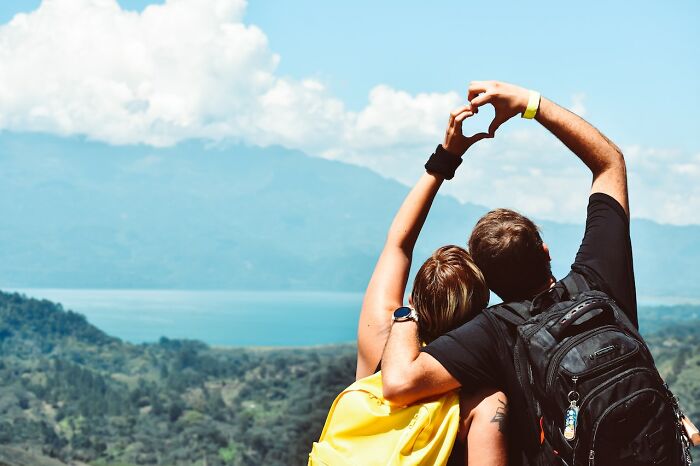 Couple makes heart with hands while overlooking a scenic lake and mountains, symbolizing travel experiences.