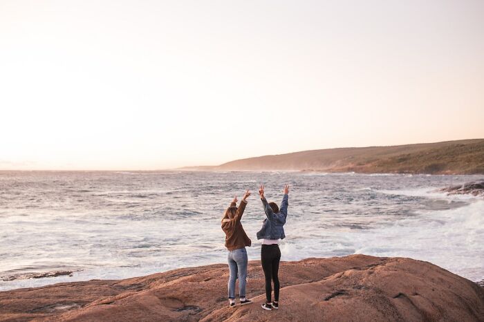 Two people standing on a rocky shore, arms raised, enjoying a breathtaking ocean view at sunset.