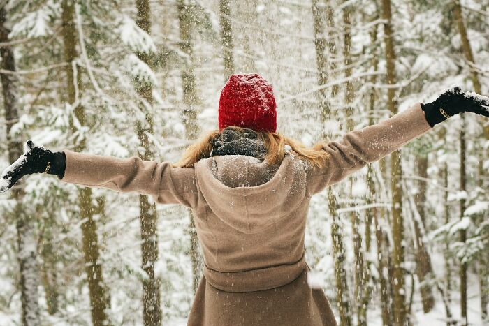Person in winter attire and red hat joyfully embracing a snowy forest scene.