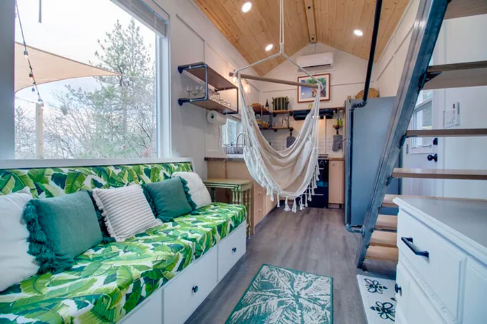 Cozy tiny house interior with a hammock, green leaf-patterned sofa, and modern kitchen under wooden ceiling.