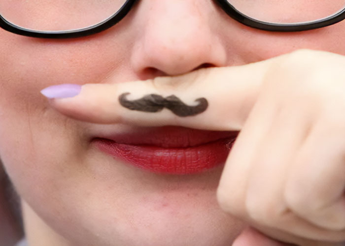 Person with mustache tattoo on finger, holding it under their nose, wearing glasses.