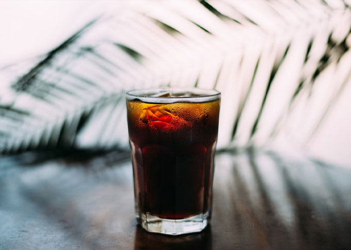 Glass of iced soda on a table with a blurred palm leaf in the background reflecting strange guest house rules.