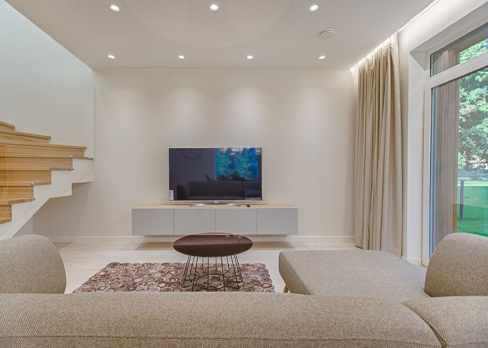 Modern living room with beige couch, round coffee table, wall-mounted TV, and large window showing outdoor greenery.