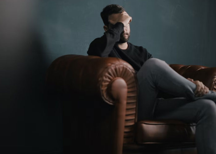 Man sitting on brown leather couch with hand on forehead, reflecting on weirdest guest house rule experiences.