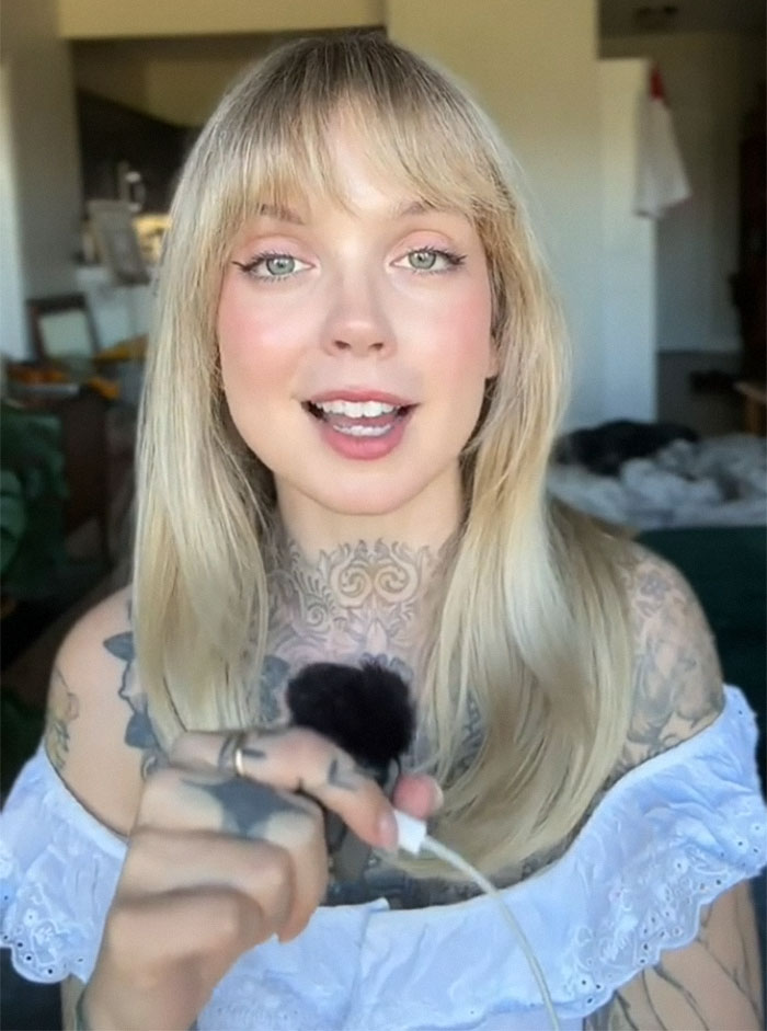 Woman with tattoos sharing regrets, holding a microphone, indoors; her tattoos are visible with long blonde hair.
