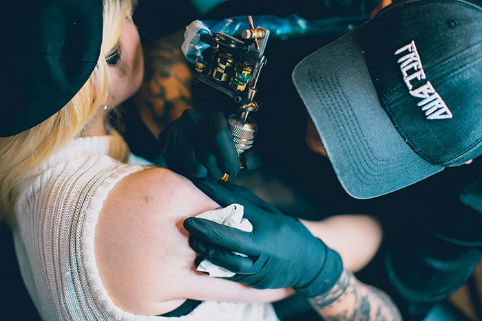 Tattoo artist working on a woman's shoulder tattoo, close-up view, black gloves and cap visible in the scene.