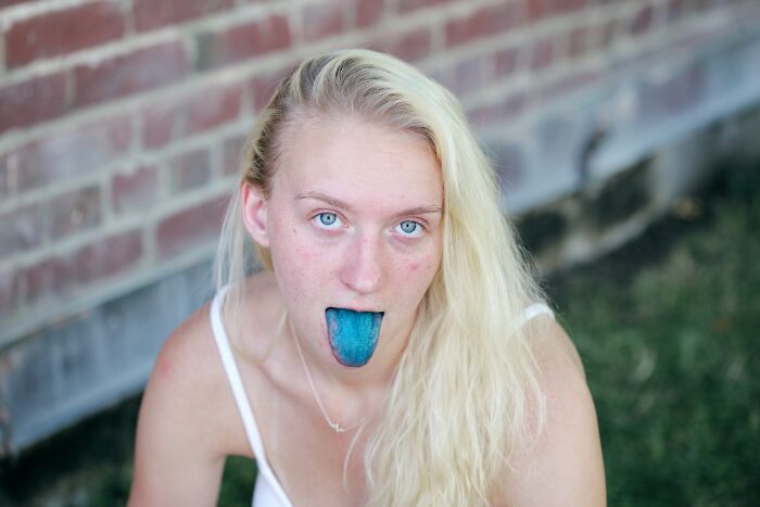 Young woman with long blonde hair showing a blue tongue, outdoors near a brick wall, illustrating the worst trend concept.