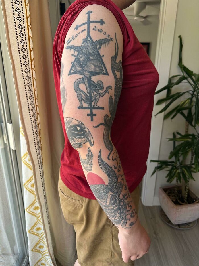 Man showcasing patchwork tattoo sleeve featuring geometric and animal designs, wearing a red shirt and tan shorts indoors.
