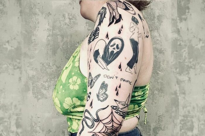 Arm with diverse patchwork tattoos, including a heart, ghost face, butterfly, and text "DON'T PANIC," wearing a green floral top.