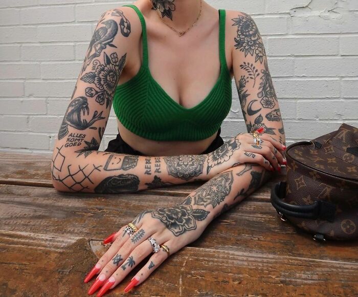 A person with patchwork tattoos on arms and neck, wearing a green top, seated at a wooden table with a bag.