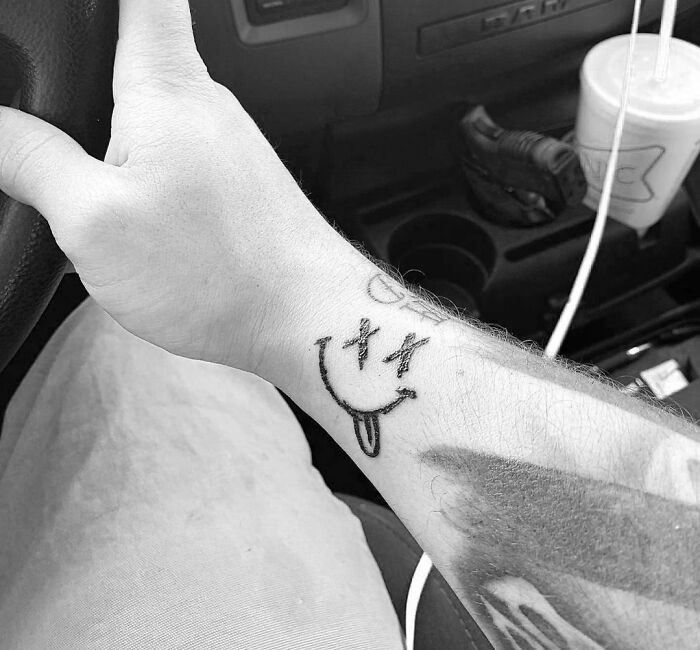 Wrist tattoo of a smiley face with X eyes and tongue out, in a car setting.