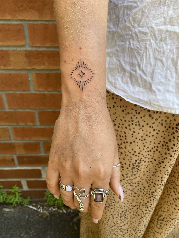 Wrist tattoo design featuring a geometric pattern, adorned hand with rings, against a brick wall background.