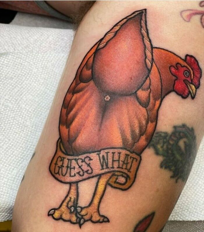 Unique, humorous tattoo of a chicken with a "Guess What" banner on a person's arm.