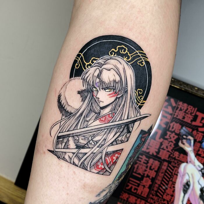 Pop culture tattoo of an anime character with long hair and a sword on an arm, framed with intricate gold designs.