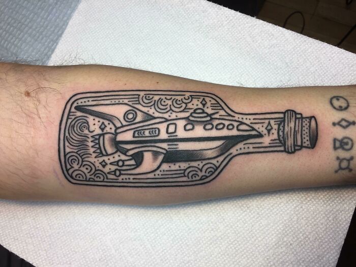 Tattoo of a spaceship inside a bottle on an arm, showcasing a unique pop culture design.