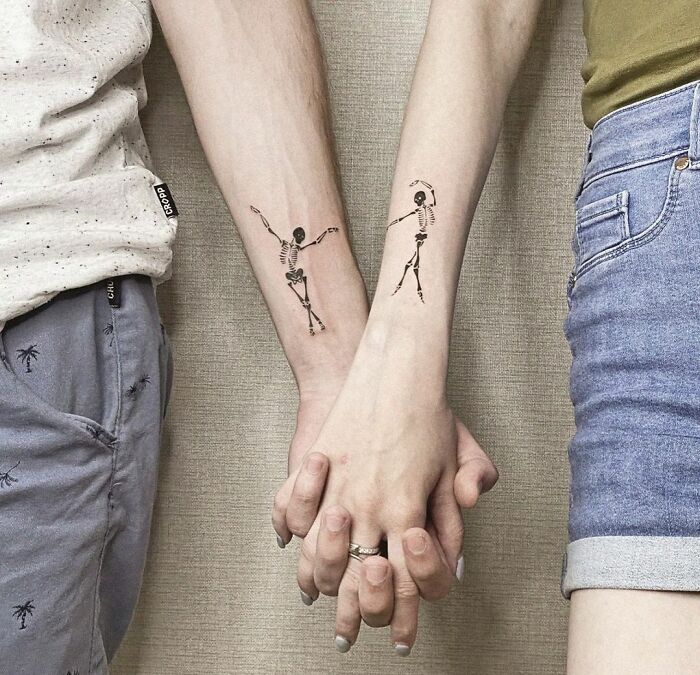 Couple holding hands with matching skeleton tattoos on their forearms, symbolizing a special connection.