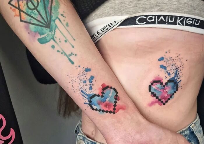 Pixelated matching heart tattoos with watercolor design on arm and stomach, showcasing special bonds.
