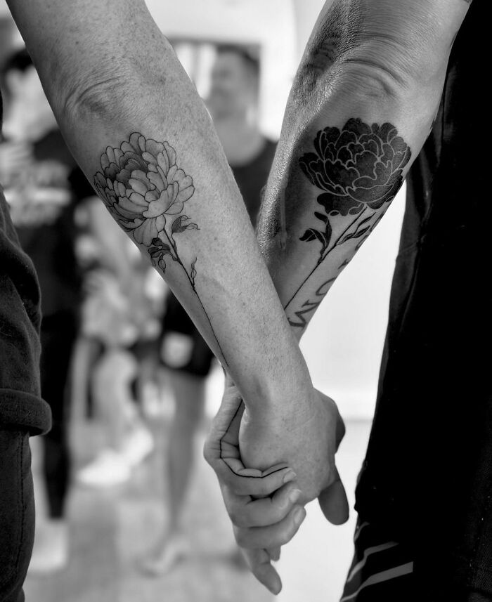 Black and white image of two people holding hands with matching floral tattoos on their forearms.