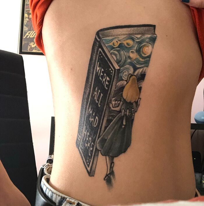 Pop culture tattoo of a girl entering a book with "We're All Mad Here" text on a person's side.