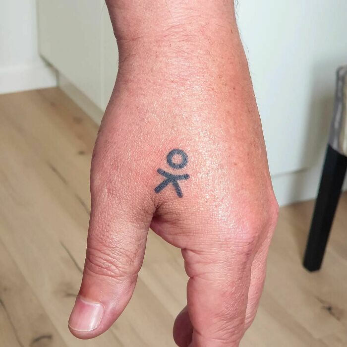Simple small hand tattoo featuring a stick figure design on the side of a thumb.