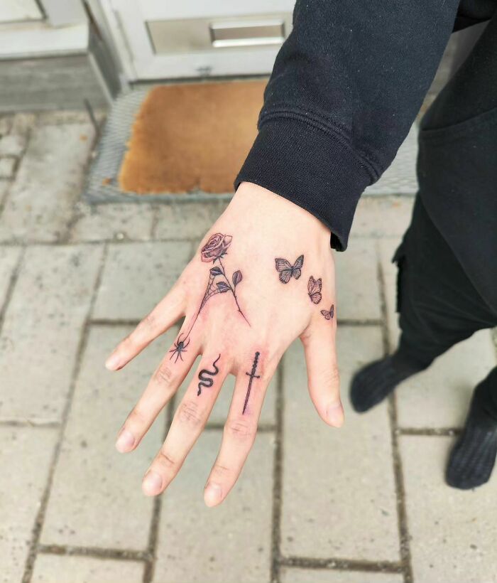 Small hand tattoos featuring a rose, butterflies, snake, spider, and cross on person's hand.