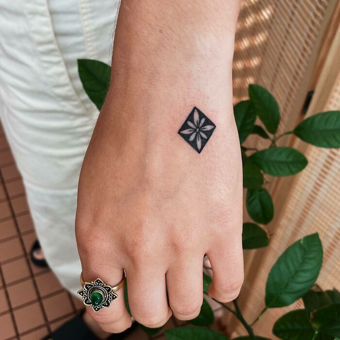 Small hand tattoo of a geometric floral design with a ring in focus, complemented by green foliage in the background.