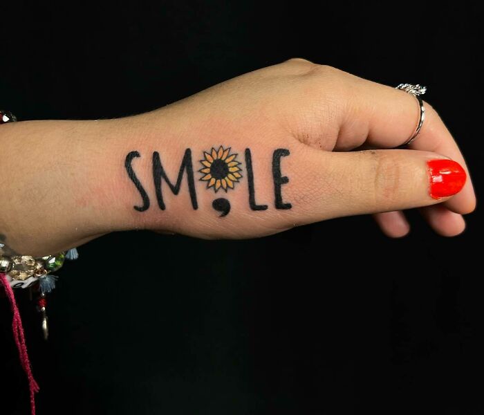 Small hand tattoo with the word "SMILE" using a sunflower for the "O" and vibrant red nail polish.