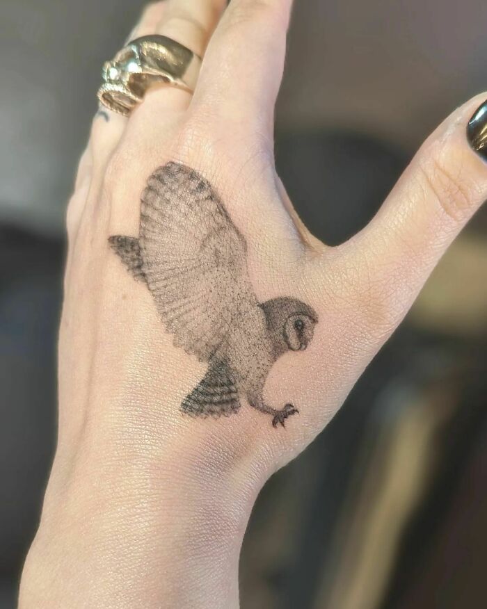 Small hand tattoo of a flying owl on a person's hand wearing a skull ring.