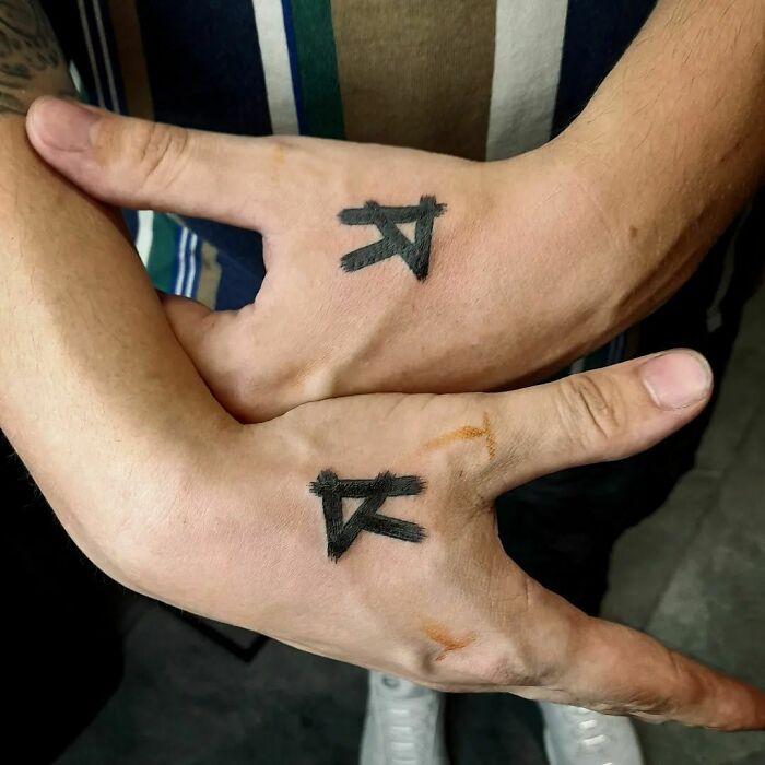 Two small hand tattoos featuring black symbols on the thumbs and webbing of intertwined hands.