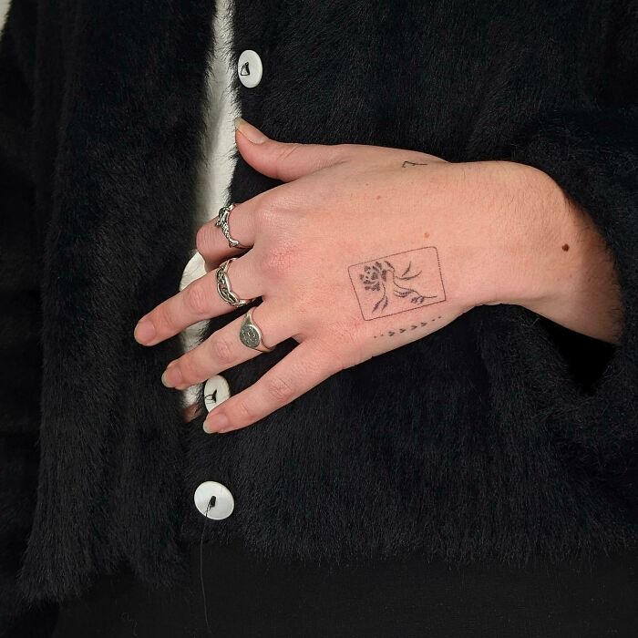 A small hand tattoo featuring a minimalist floral design with silver rings on fingers, against a black fabric background.