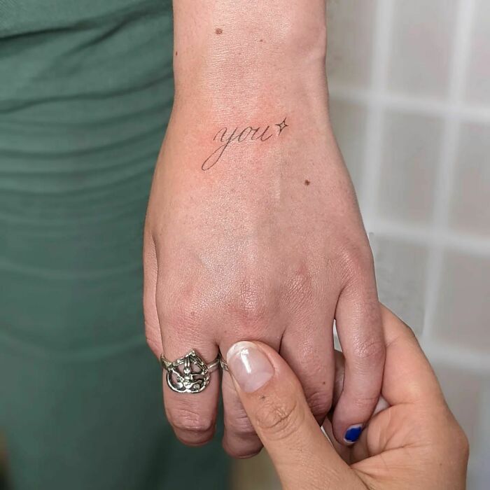 Small hand tattoo with the word "you" and a star, held by another hand with blue nail polish.