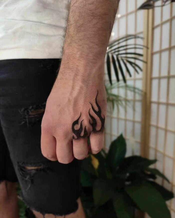 Man's hand with small flame tattoo, set against a plant-filled background, showcasing unique small hand tattoo design.