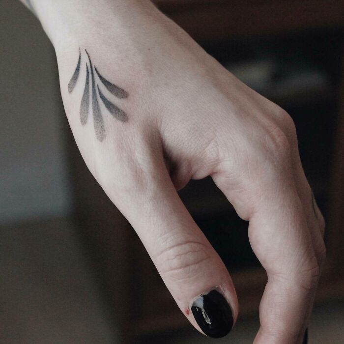 Black and gray small hand tattoo with abstract design on the back of the hand, black nail polish visible.