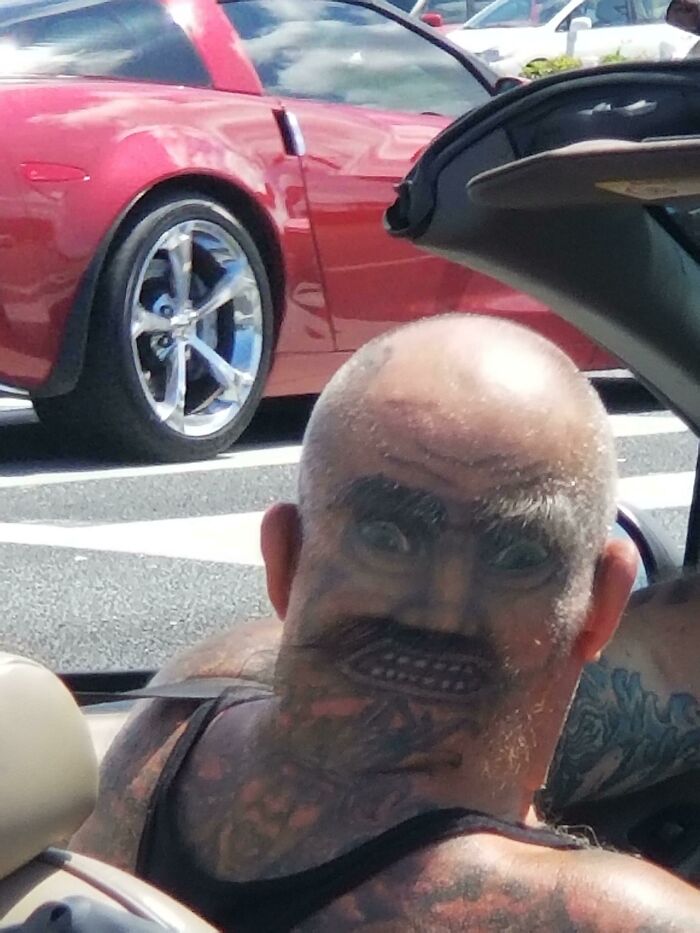 Man with a unique tattooed face design on the back of his head sitting in a convertible car.