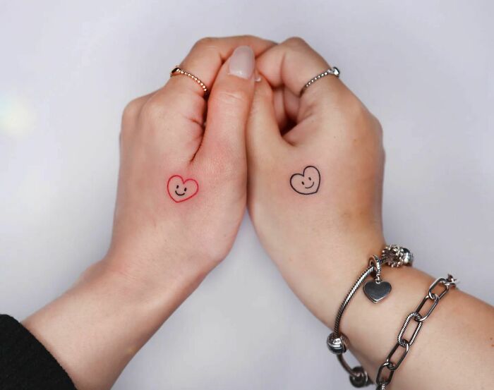 Two hands with small heart tattoos featuring smiley faces, surrounded by silver jewelry.