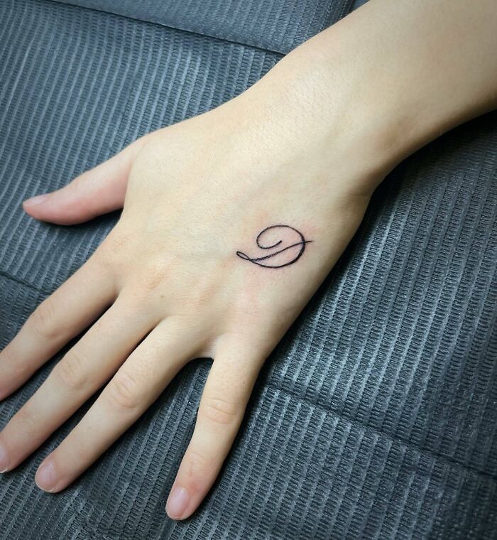 Elegant small hand tattoo of a cursive letter "D" on a person's hand.