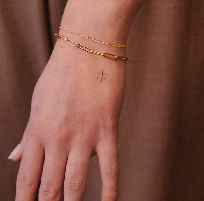 Small hand tattoo of a minimalist smiley face on wrist, alongside gold chain bracelets.