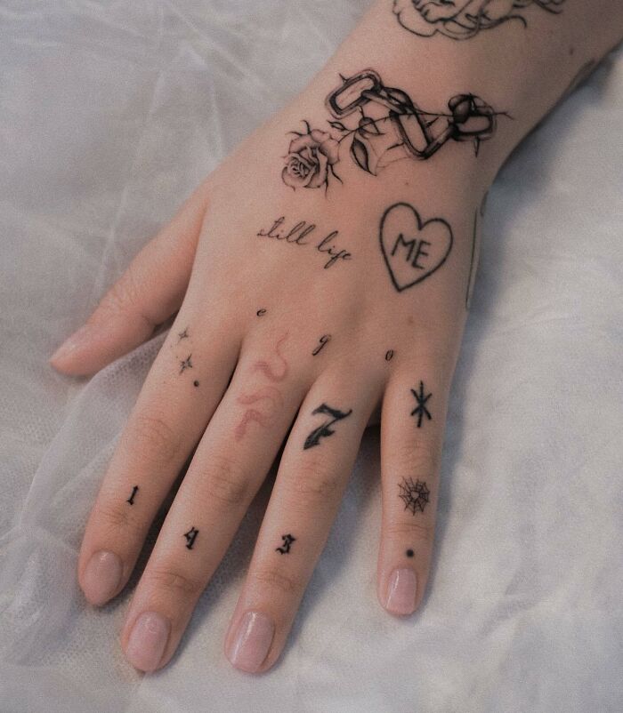 Hand adorned with small tattoos, including a heart, numbers, and a rose, showcasing small hand tattoo designs.