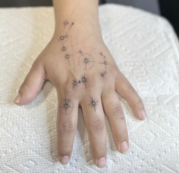 Small hand tattoo with constellation design on a person's hand seen on a textured white background.