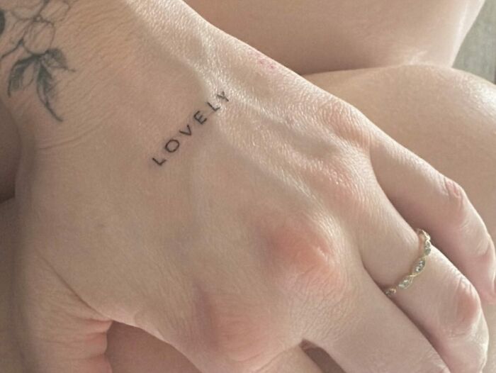 Small hand tattoo reading "Lovely" with a floral design, showcasing delicate art in a minimalist style.