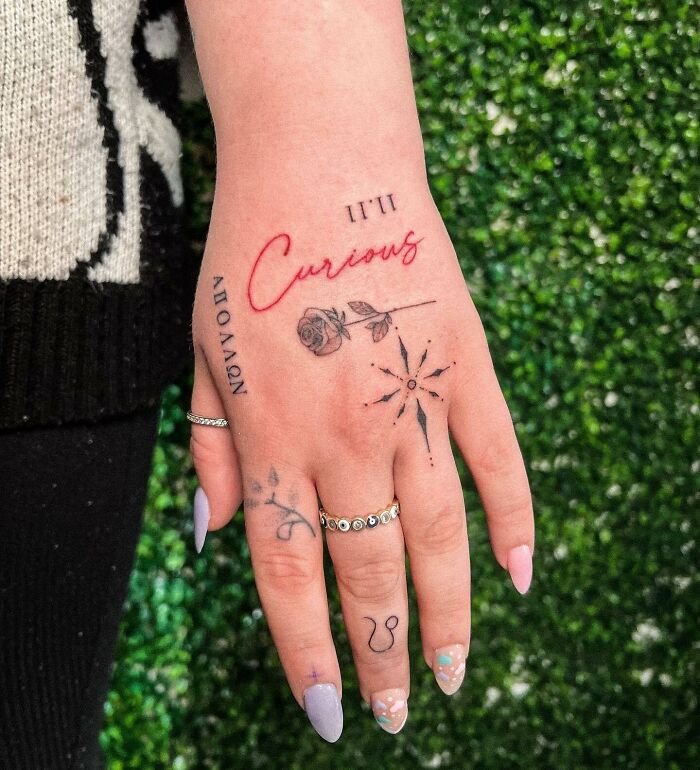 Small hand tattoos featuring cursive text, a rose, and geometric designs on a woman's manicured hand.