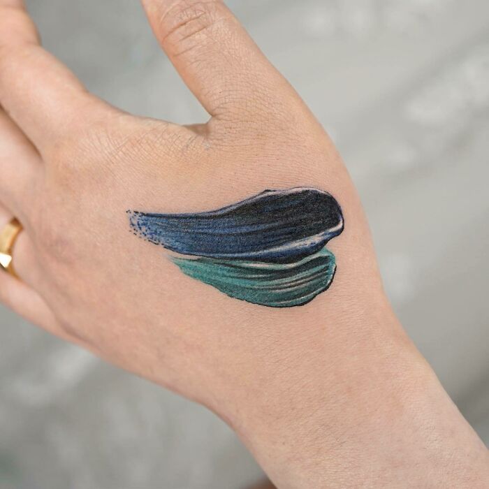 Small hand tattoo of blue and green paint strokes on the back of a hand.